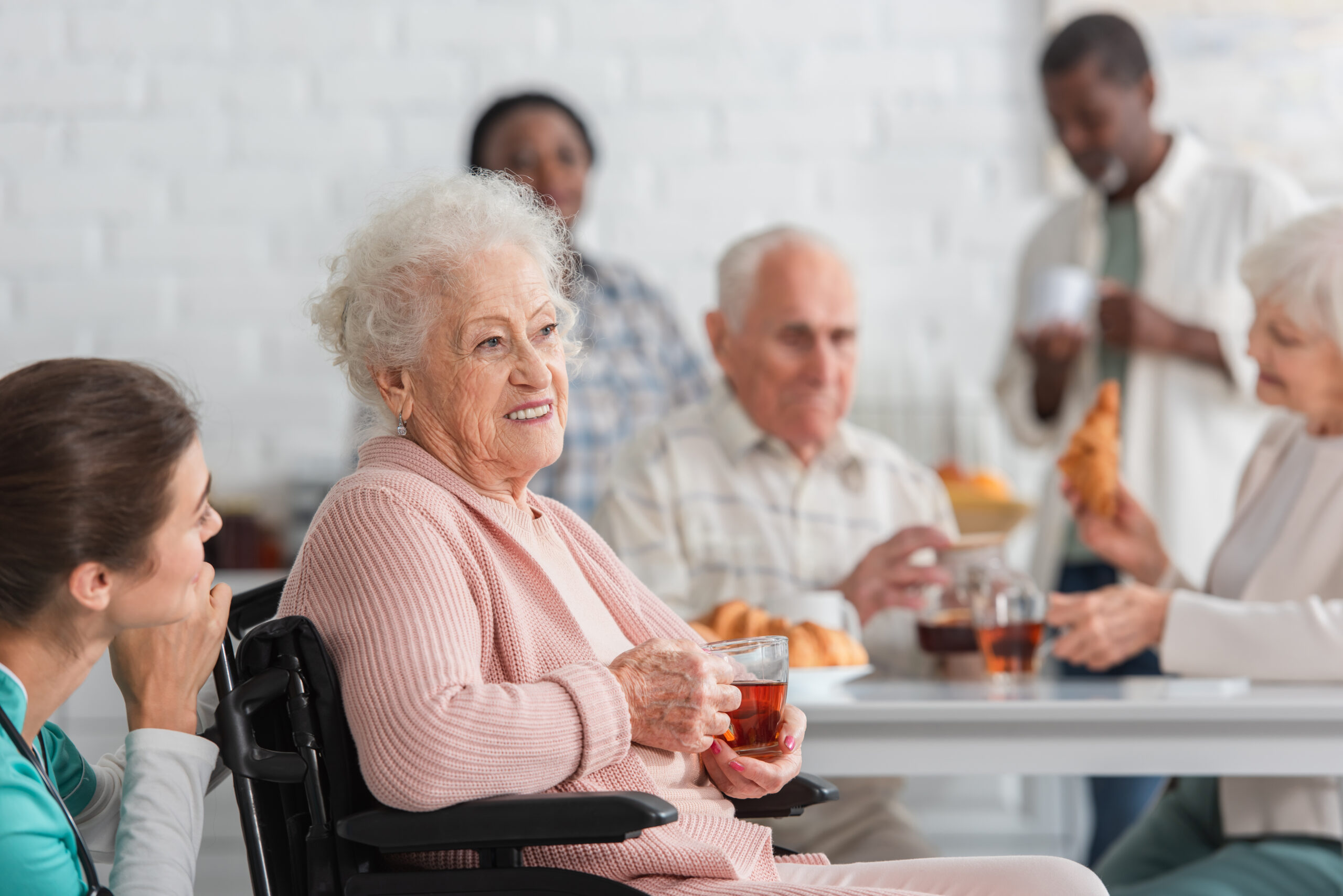 Senior woman holding tea in wheelchair near nurse and blurred in